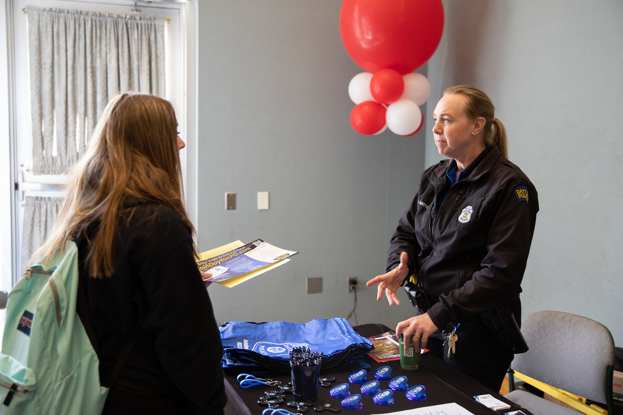 Uniformed officer speaks with a visitor at an event registration desk, with lanyards and brochures on the table and red and white balloons in the background.