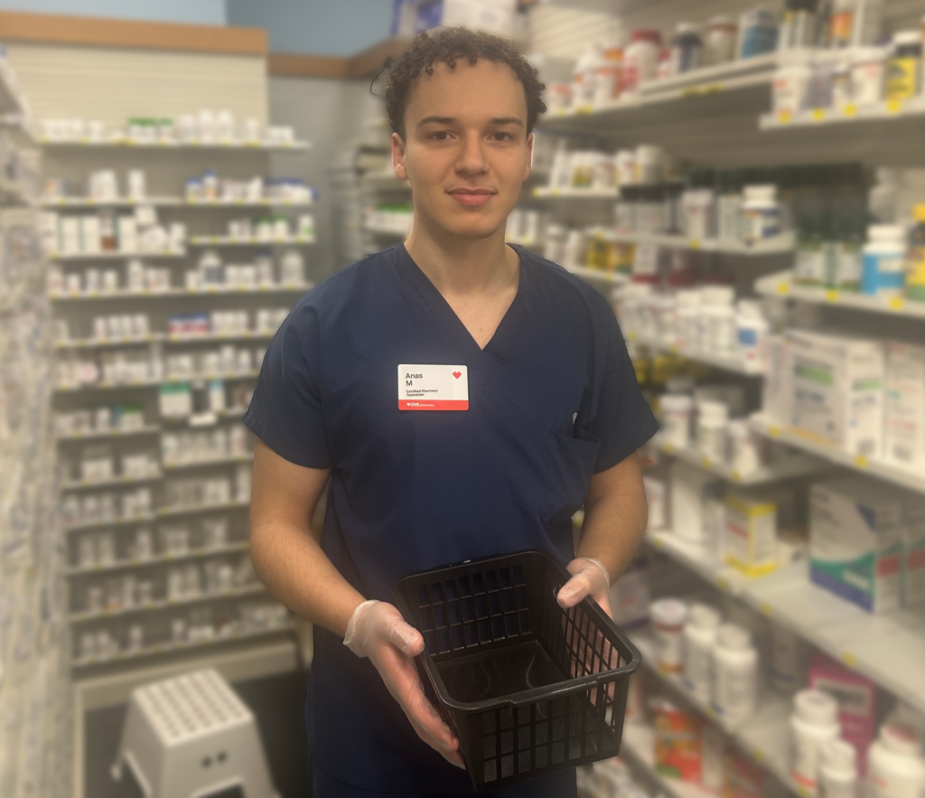 Pharmacist in navy scrubs holding a black plastic basket in a pharmacy aisle with shelves of medicines in the background.