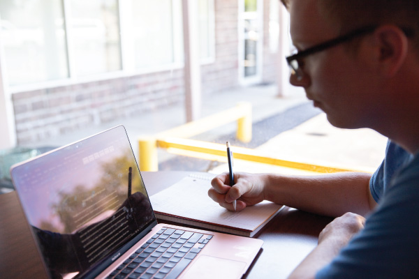 A Communication Studies student looking at his laptop and writing with a pen on a piece of paper.
