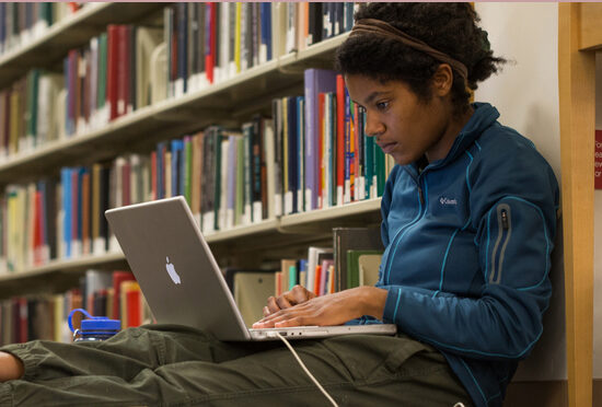 Student working on computer in front of book stacks at the library.
