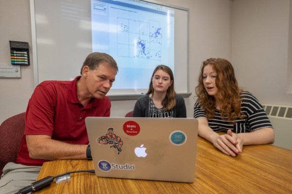 Three people sit around a wooden table in a classroom, looking at a laptop with a world map projected on a screen behind them.
