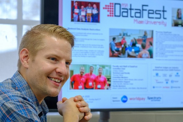Smiling man in a blue checkered shirt sits in front of a display screen labeled 'DataFest' at Miami University.