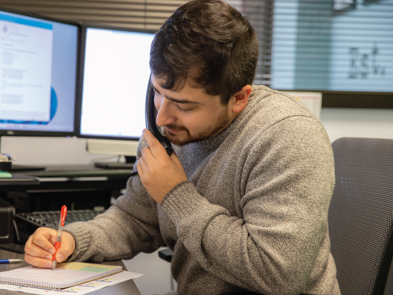 Man in a gray sweater sits at a desk, talking on a phone while writing in a notebook with a red pen on a cluttered desk.