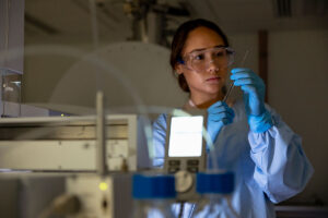 female student wearing a blue lab coat, working in lab with safety goggles and machinery