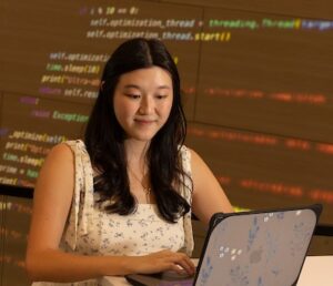student working at laptop with html code on wall behind her