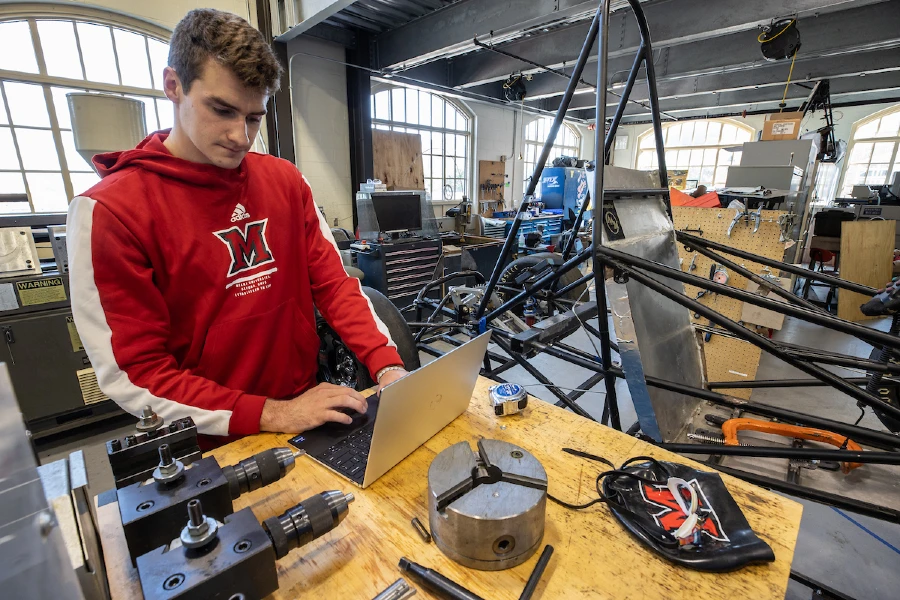 student wearing a miami sweatshirt, working on laptop in engineering lab, with a car in background