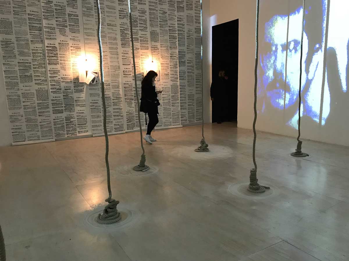 Contemporary art space with hanging ropes, a wall covered in printed pages, and a projected portrait of a man on a white wall.