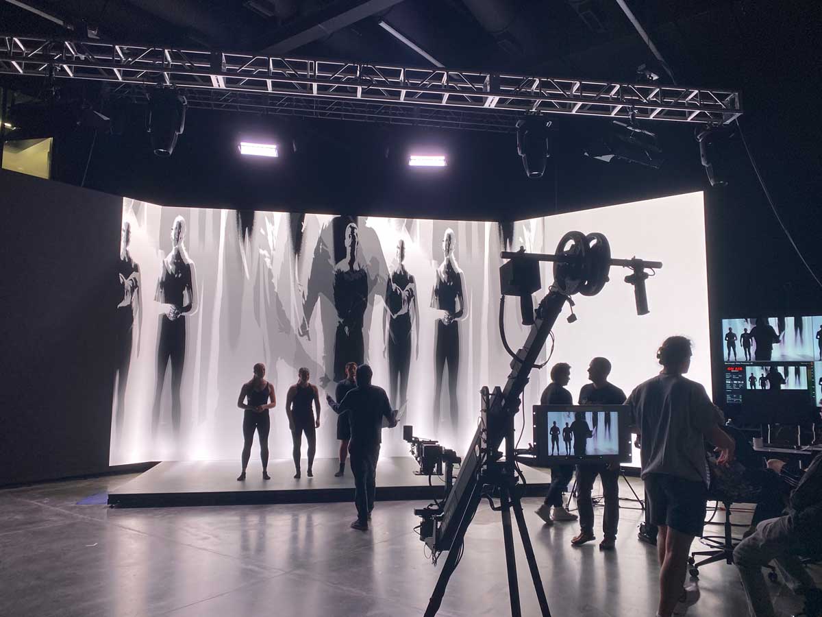 Studio production with performers, crew, and a large digital backdrop showing black-and-white figures.
