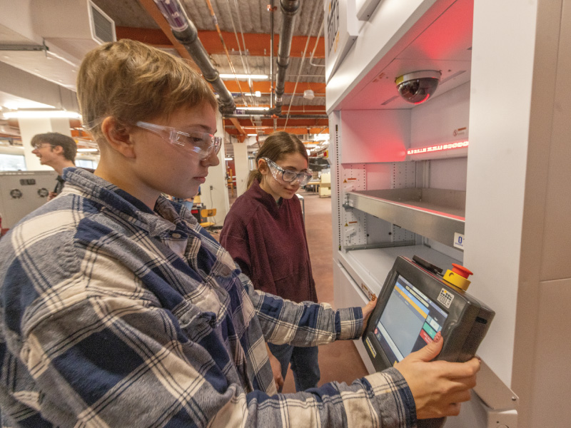 Two students wearing safety goggles adjust a touchscreen control panel on a large lab machine in an industrial classroom or workshop environment.