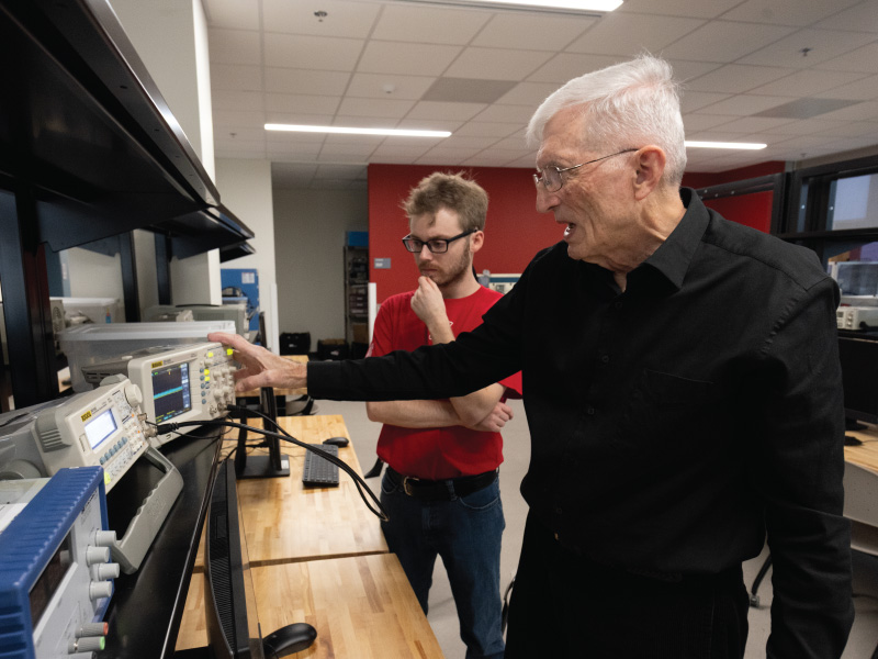 Two men in a lab look at electronic test equipment; the older man in black points to a device while the younger man in red observes thoughtfully.