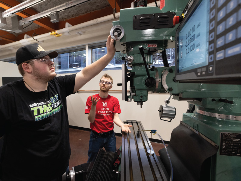 Two students operate a large green milling machine in a workshop; one adjusts a control wheel while the other talks and gestures.