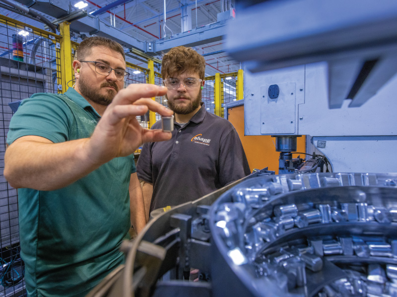 Two male technicians wearing safety glasses inspect a small metal part held up to a large industrial machine in a factory setting