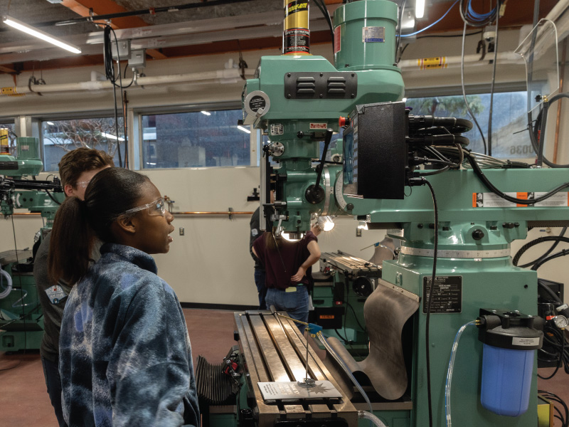 Three students wearing safety goggles stand beside a large green vertical milling machine in a machining lab, examining the metal workpiece on the table.