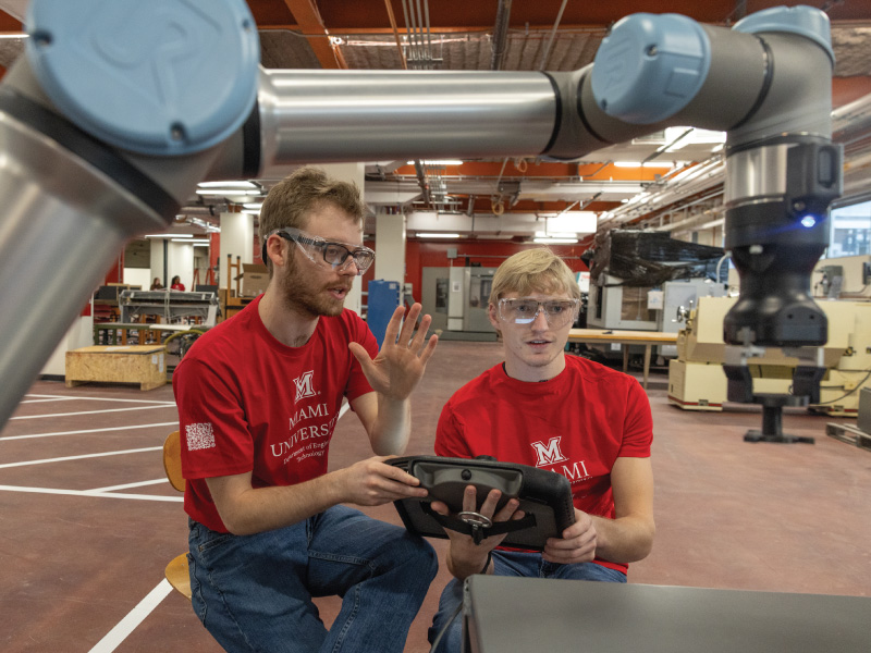 2 Engineering Technology students looking at a robot arm and holding a tablet used to control the robot.