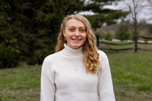 Young woman with long blonde hair wearing a white turtleneck sweater, smiling outdoors in a grassy area with a wooden fence in the background.